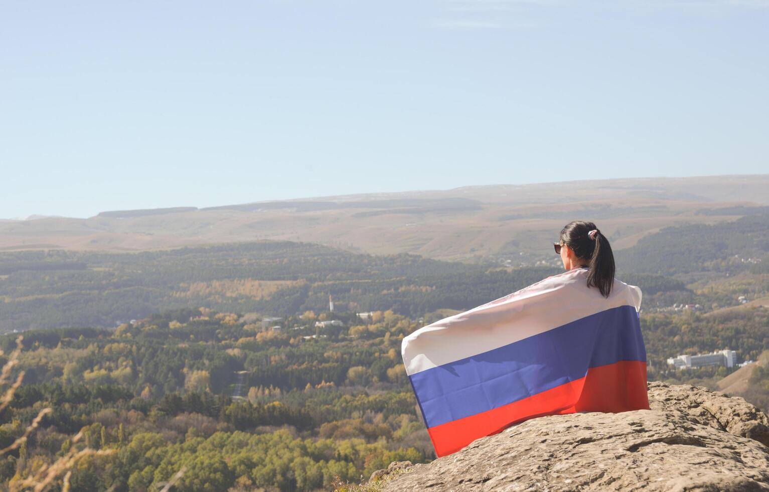 A young brunette woman sits on a stone and looks at the mountains around, her shoulders are covered with a Russian flag photo