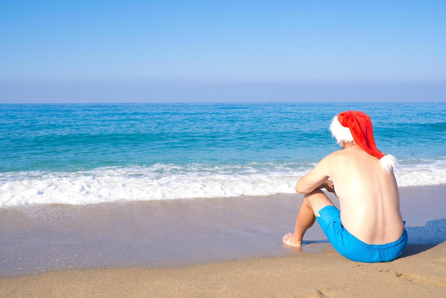 A man in a Santa Claus hat sits on the seashore, rear view. Christmas Santa Claus relaxing on the ocean tropical beach sand - New Year travel destinations, holidays in hot countries, travel photo