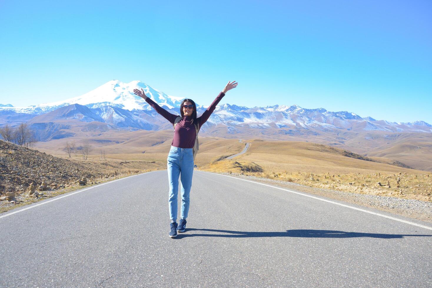 A happy female traveler with a backpack behind her back and her arms raised up rejoices at her trip to the mountains. Elbrus on a clear autumn day. View of Elbrus, North Caucasus, Russia photo