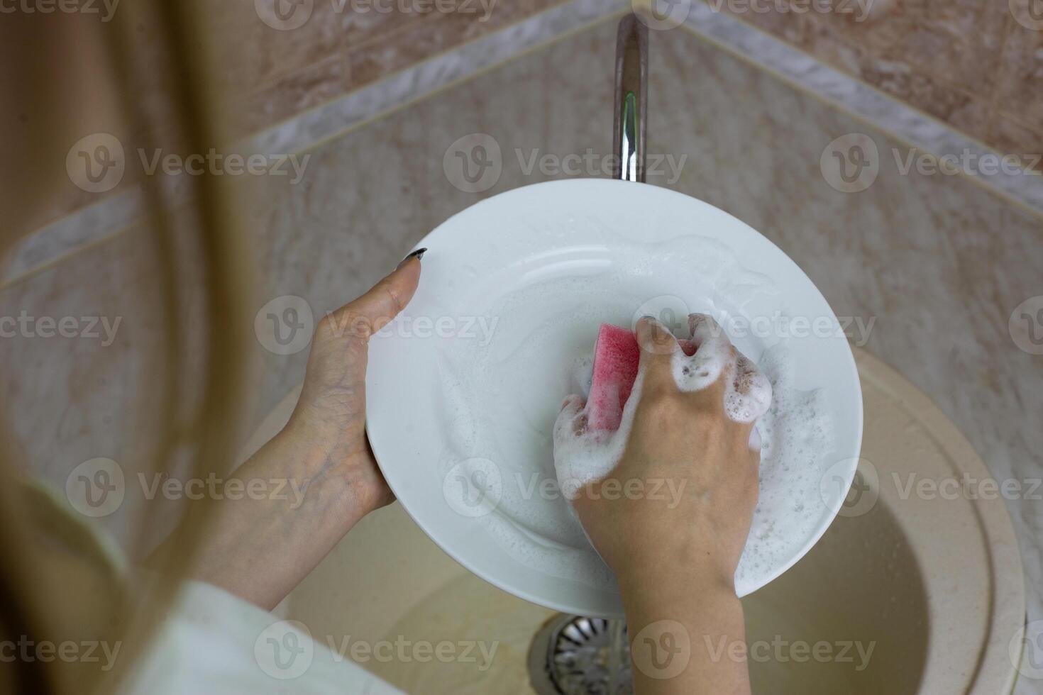Female hands washing white plate in sink with sponge and foam from detergent in the kitchen at home. Cleaning dishes after meals, daily routine in apartment photo