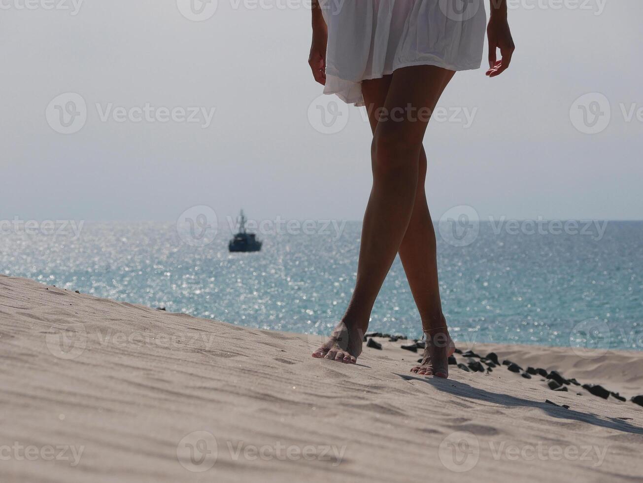 A barefoot girl wearing white dress walks along the sand dune against the backdrop of the sea on Sardinia beach. Cropped photo, legs close up photo