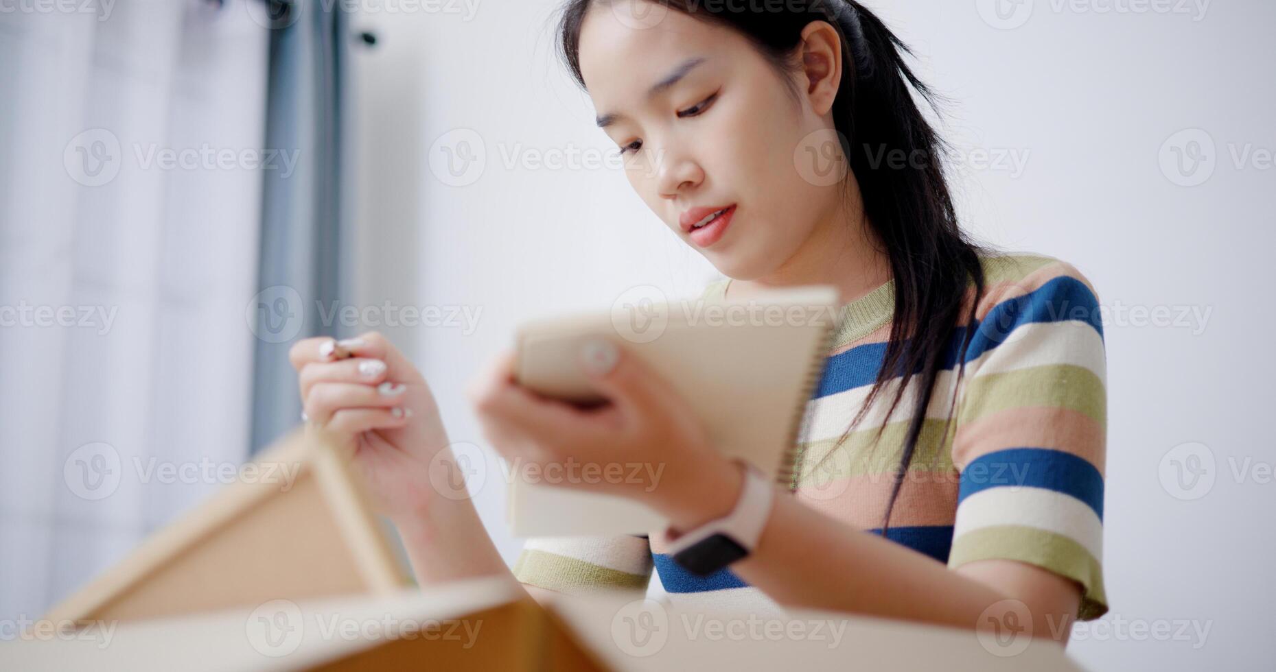 Low angle view, Young Asian woman is taking notes while checking belongings in cardboard boxes in preparation for moving to a new house. Moving house and lifestyle concept. photo