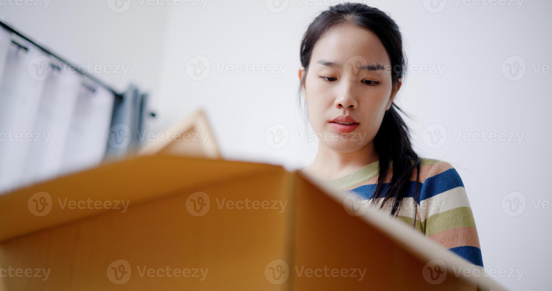Low angle view, Young Asian woman is taking notes while checking belongings in cardboard boxes in preparation for moving to a new house. Moving house and lifestyle concept. photo