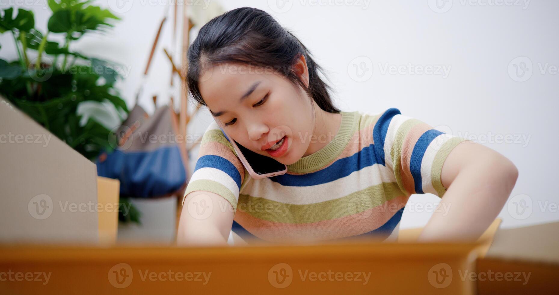 Portrait of Young Asian woman talking on mobile phone while while packing belongings into cardboard boxes to prepare for a move. Moving house and lifestyle concept. photo