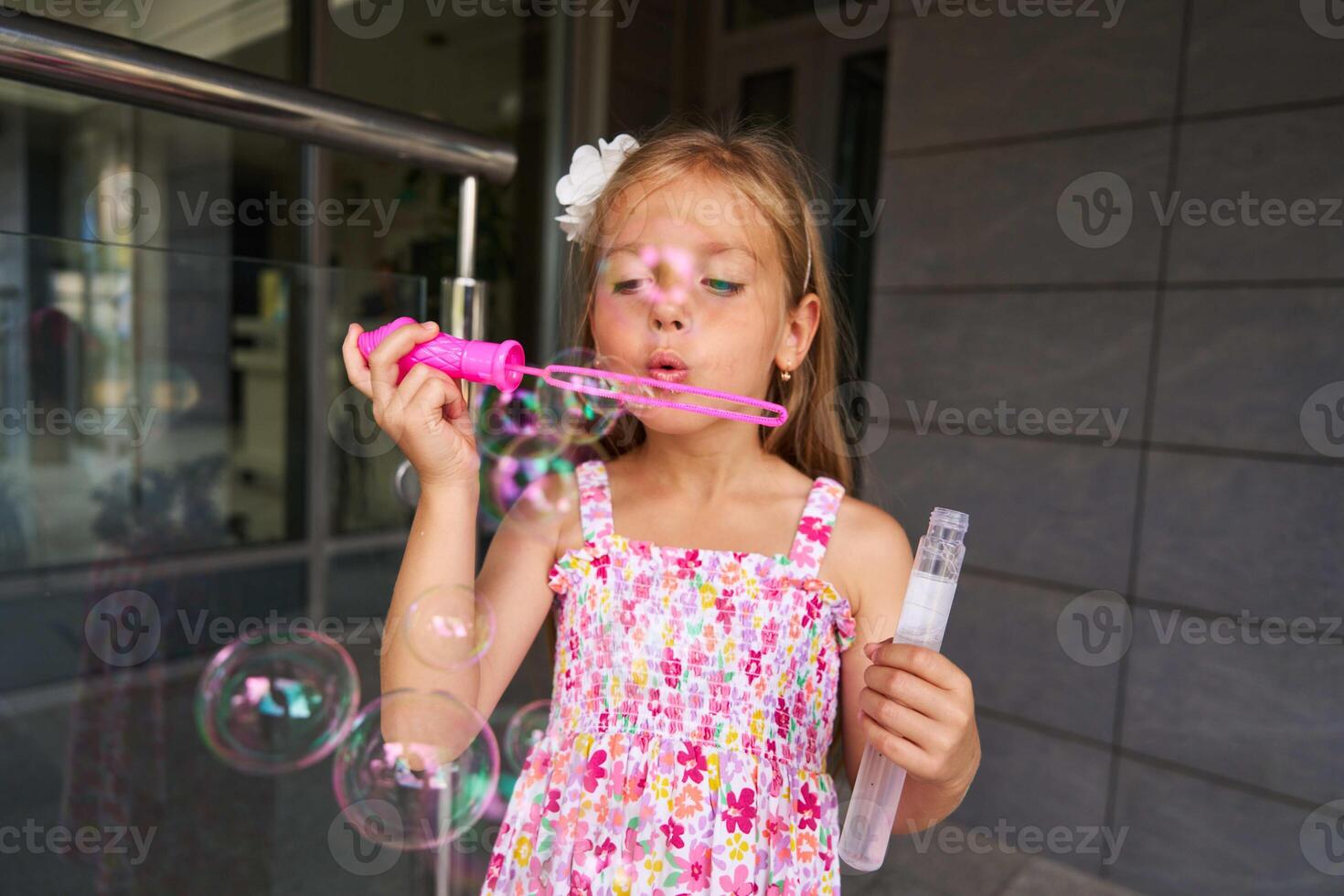 Child Blowing Colorful Bubbles With Pink Wand on Sunny Day in Urban Setting photo