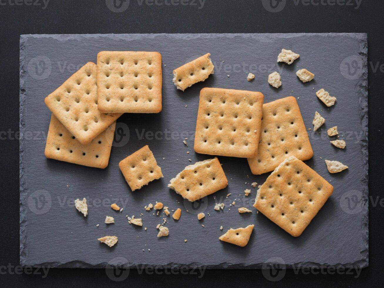 Square crackers with pieces and crumbs on slate gray background. Dry salt cracker cookies with fiber and dry spices. Top view or flat lay. photo