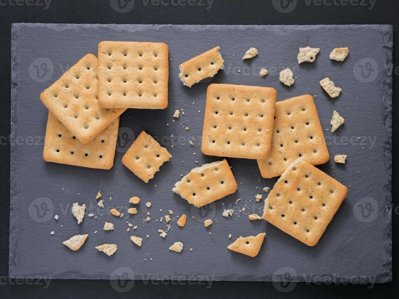 Square crackers with pieces and crumbs on slate gray background. Dry salt cracker cookies with fiber and dry spices. Top view or flat lay. photo