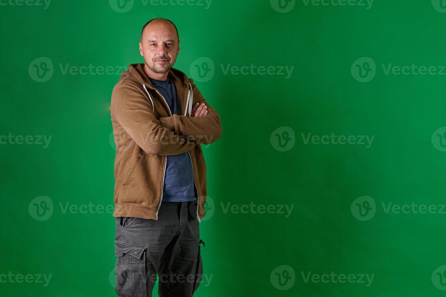 Middle aged bald man in casual attire with crossed arms poses confidently against a green studio backdrop. photo