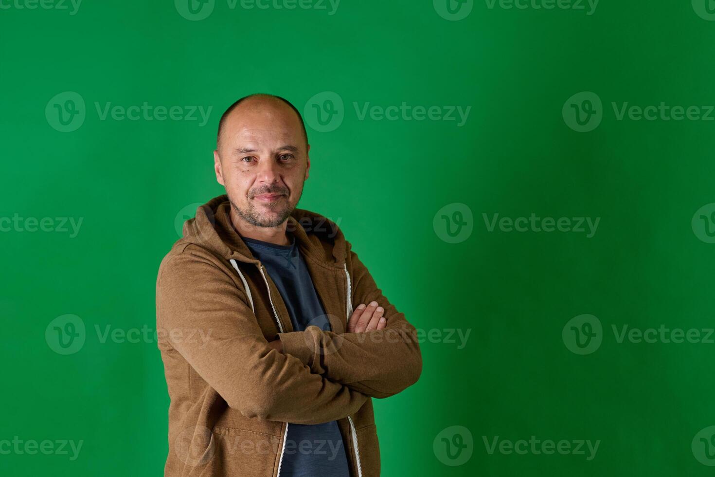 Middle aged bald man in casual attire with crossed arms poses confidently against a green studio backdrop. photo