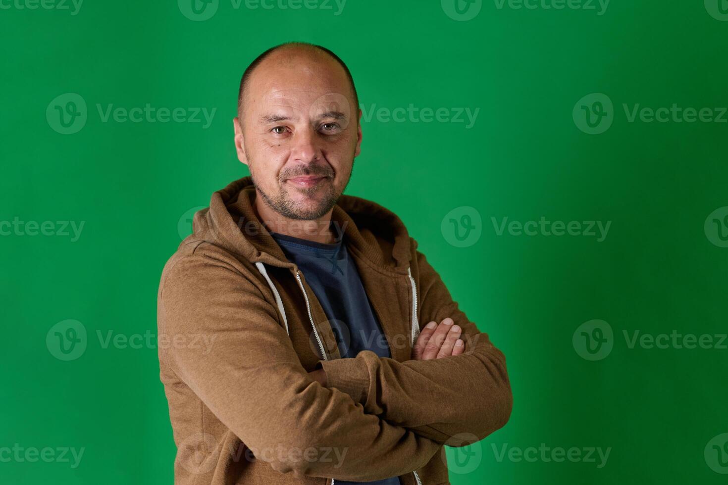 Middle aged bald man in casual attire with crossed arms poses confidently against a green studio backdrop. photo