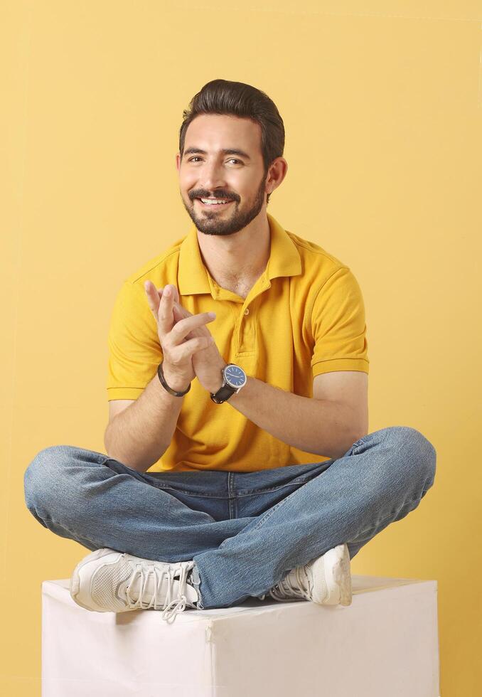 Portrait of a young adult stylish beard male model in yellow floral shirt and jeans with smile full height and sitting studio shots photo