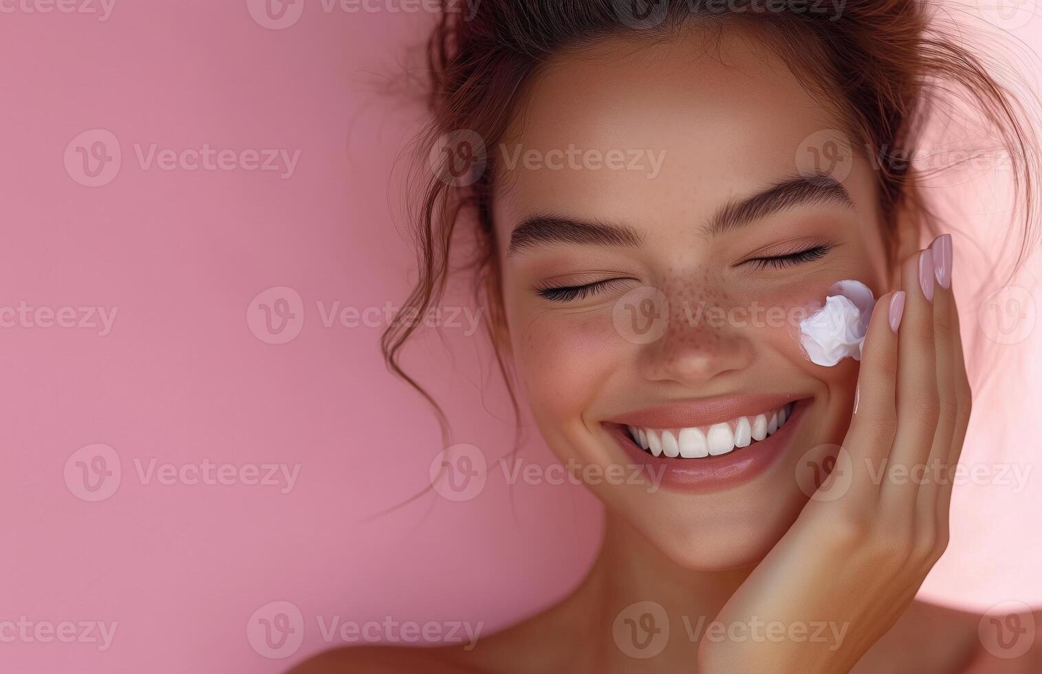A lovely woman with closed eyes smiles while applying cream to her cheek with one hand photo
