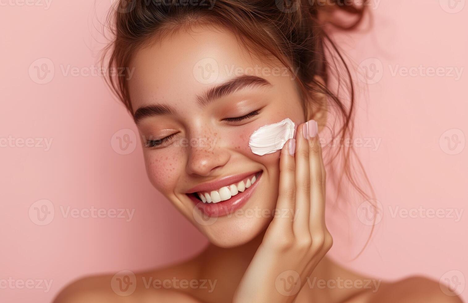 A lovely woman with closed eyes smiles while applying cream to her cheek with one hand photo