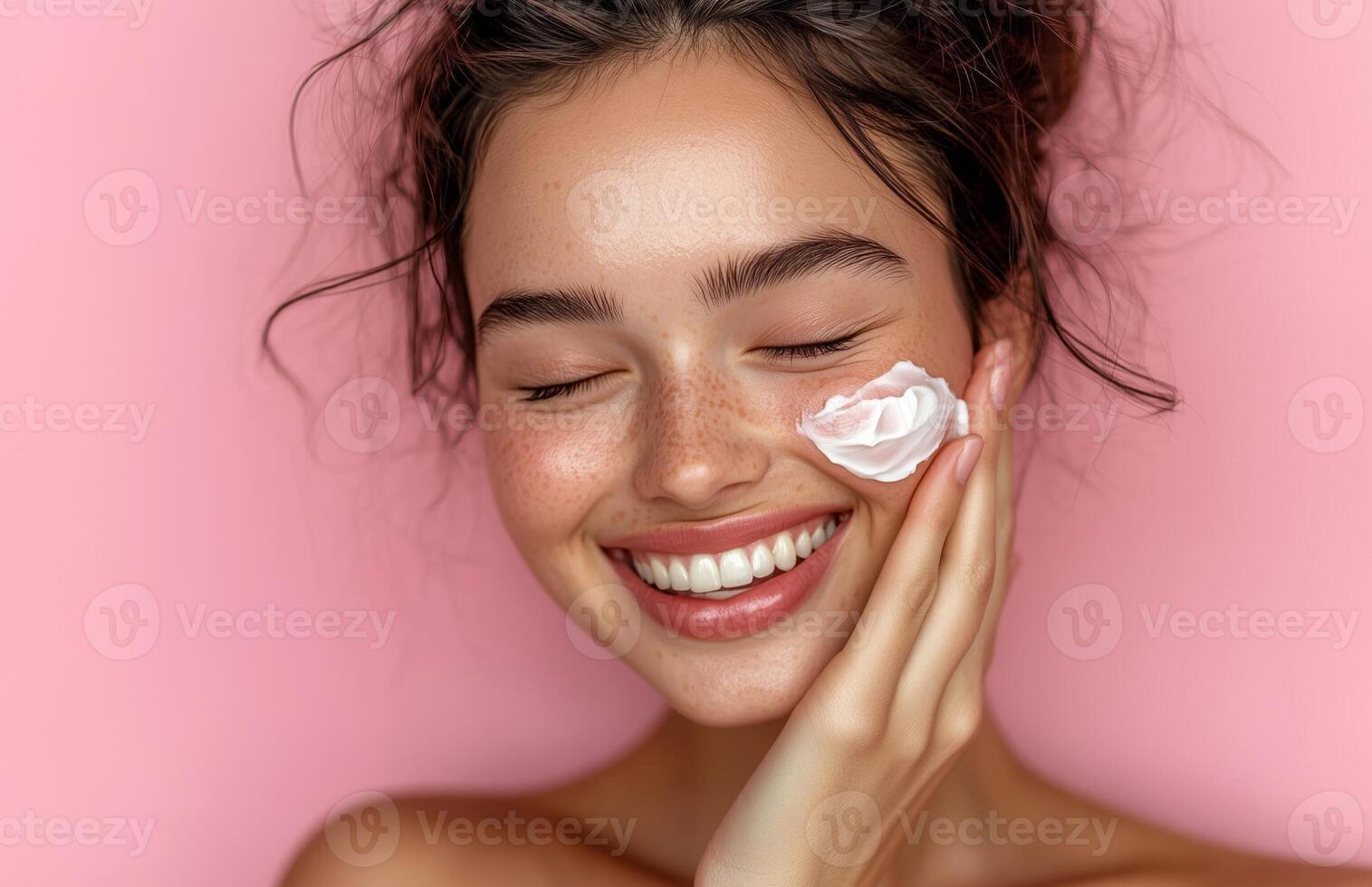 A lovely woman with closed eyes smiles while applying cream to her cheek with one hand photo