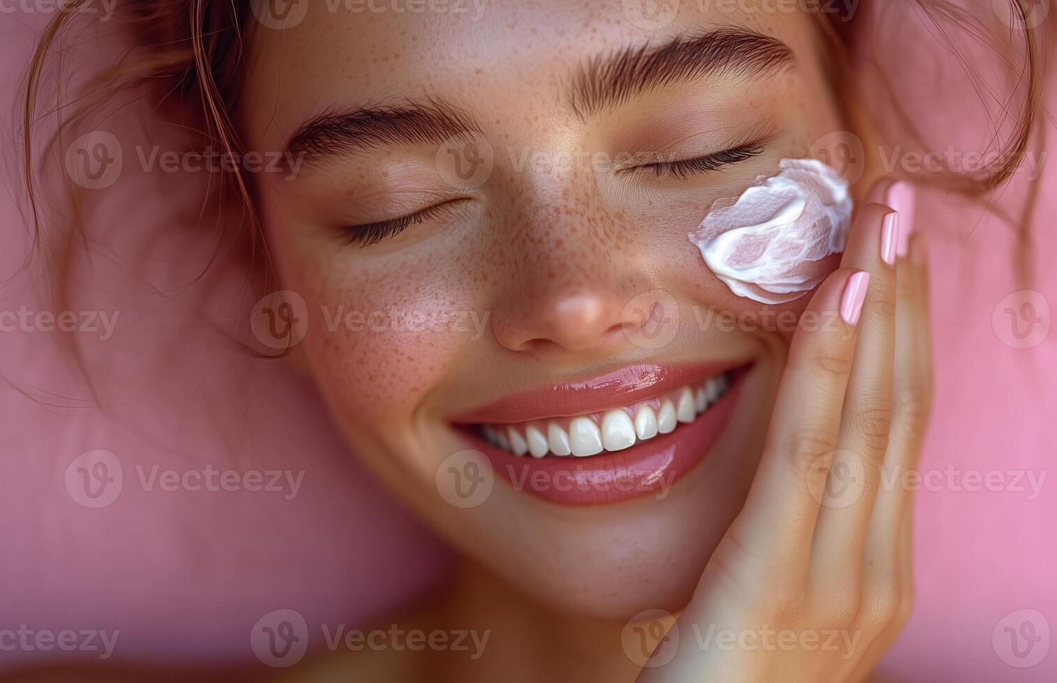 A lovely woman with closed eyes smiles while applying cream to her cheek with one hand photo