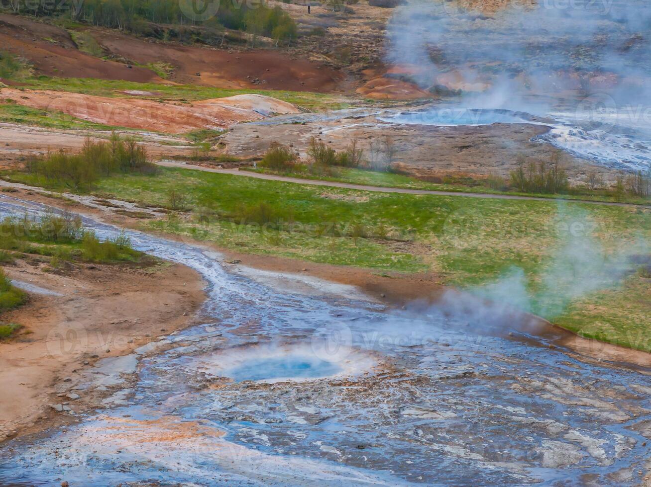 A stunning aerial view of a geothermal area in Iceland, featuring a steaming hot spring with a blue pool. The landscape includes brown, green, and blue hues with a flowing stream. photo