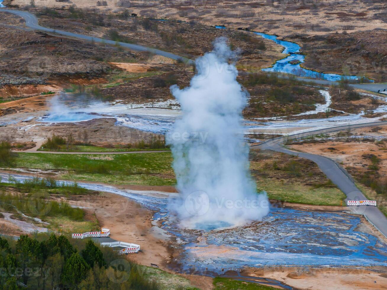 An aerial shot capturing a geyser erupting in Iceland, surrounded by a muddy area and a small stream. A winding road and a small forest are also visible. photo