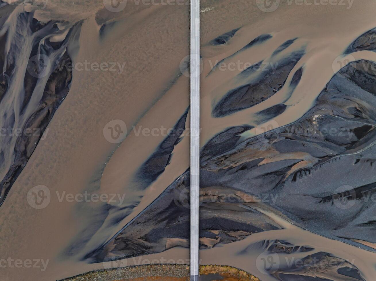 A stunning aerial view of a light brown river winding through dark gray and brown rock in Iceland, split by a white road or bridge, creating an abstract pattern. photo