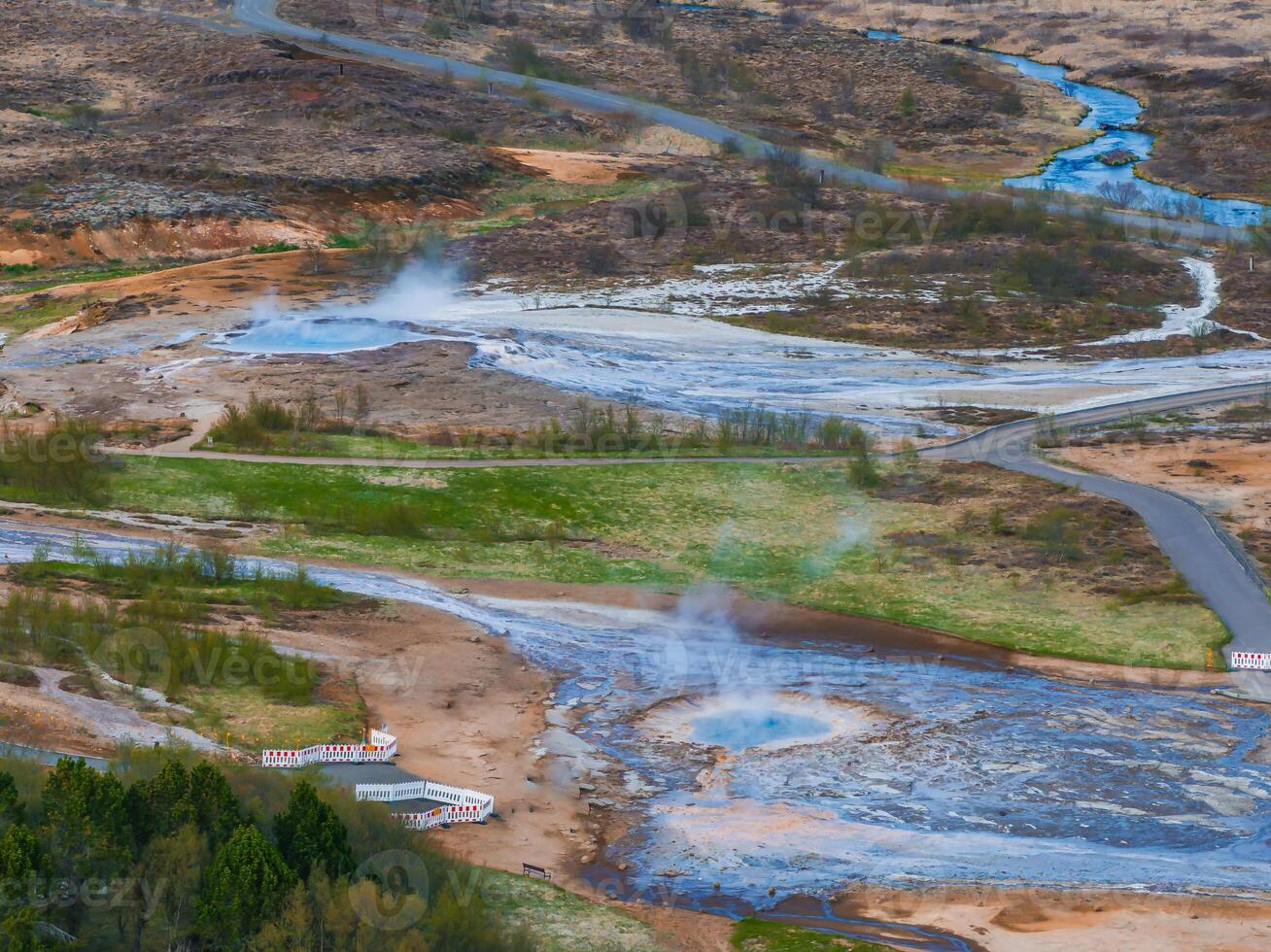A stunning aerial view of two vibrant blue geysers surrounded by a barren landscape with a winding river in Iceland. The mix of brown, green, and blue creates a striking contrast. photo