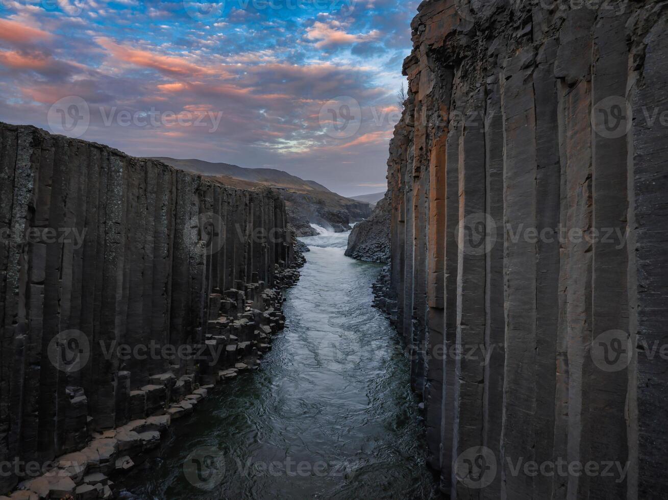 A stunning aerial view of a river winding through a narrow canyon with tall, dark rock formations. The sky above features a beautiful mix of blue and pink clouds. photo