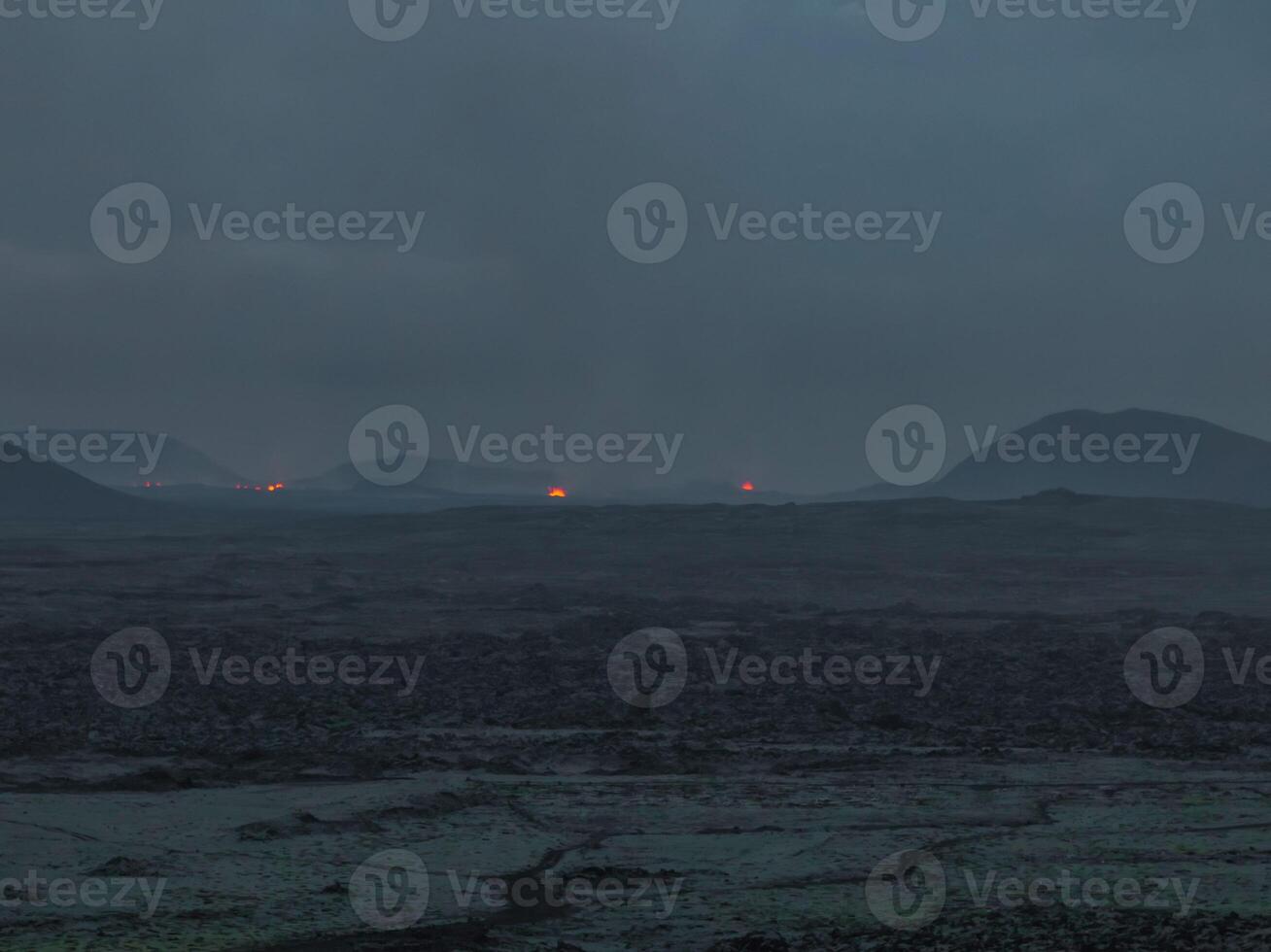 Aerial view of a dark volcanic landscape in Iceland at dusk, featuring small glowing red craters. The sky is a deep blue, contrasting with the black ground. photo