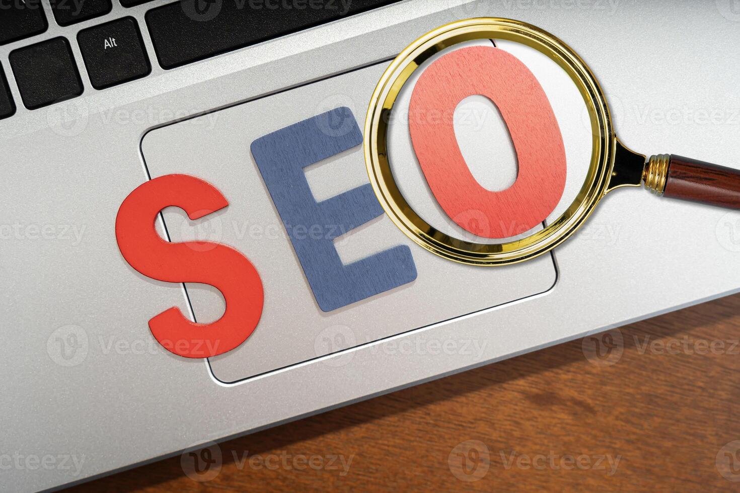 Magnifying glass magnifying wooden letters stands for SEO abbreviation lying on laptop at desk photo