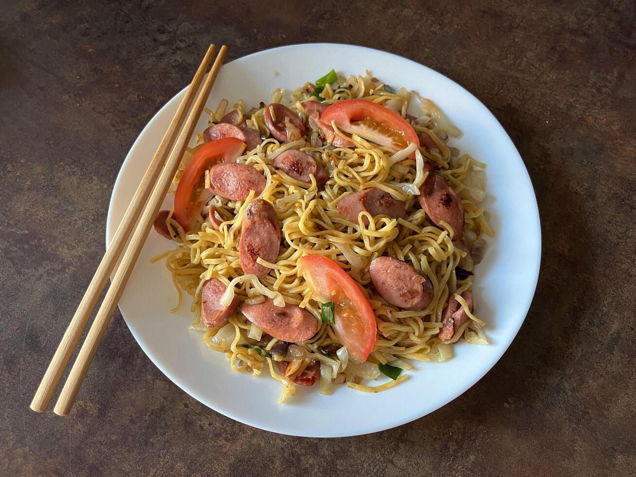 Mie goreng sosis or sausage fried noodles is an Indonesian noodle dish cooked with a sticky, savoury sweet sauce, sausage, and Chinese cabbage served in white plate on dark background. photo