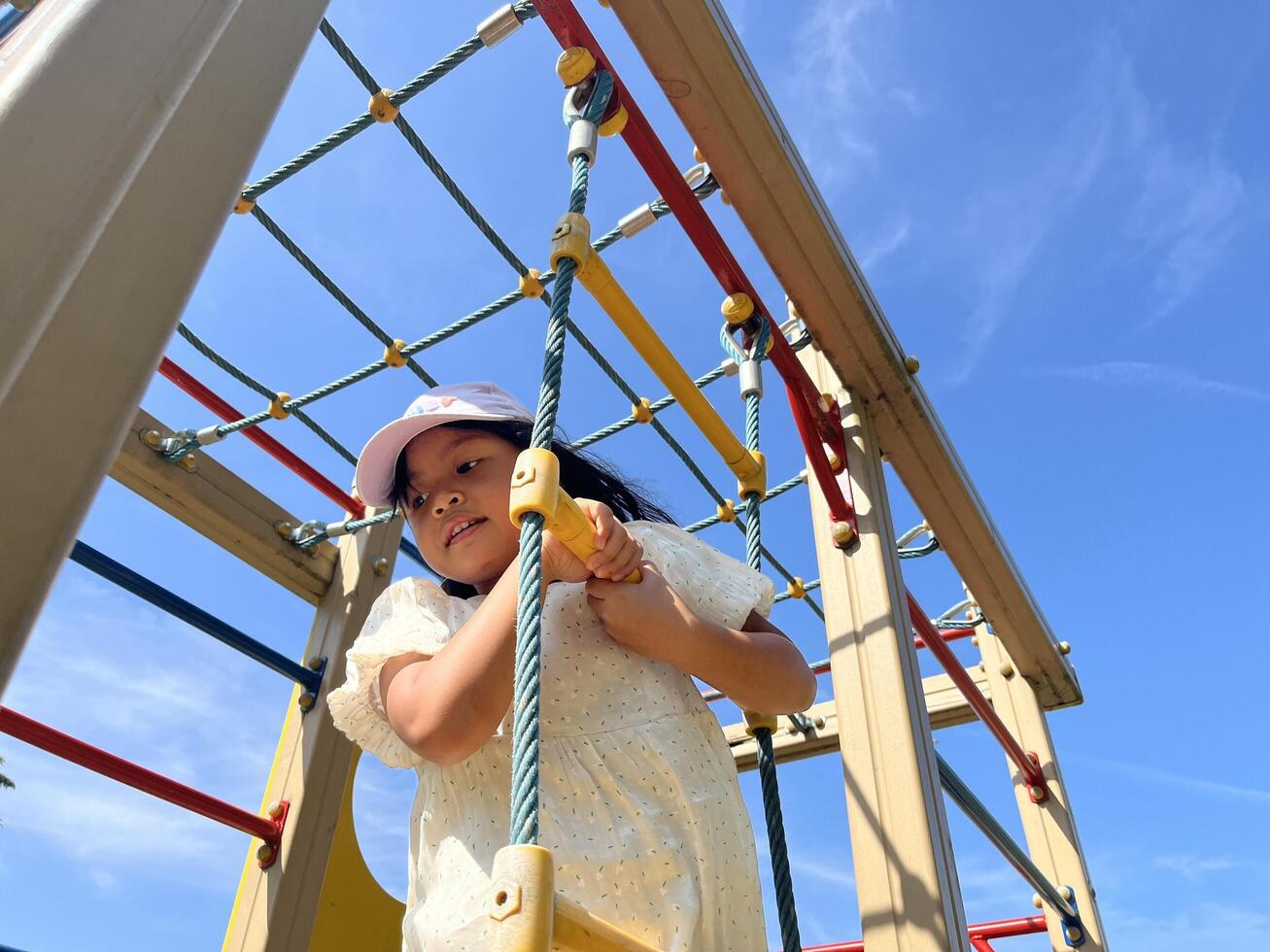 Happy Asian little girl playing rope climbing in playground in summer sunny day. Bottom view photo