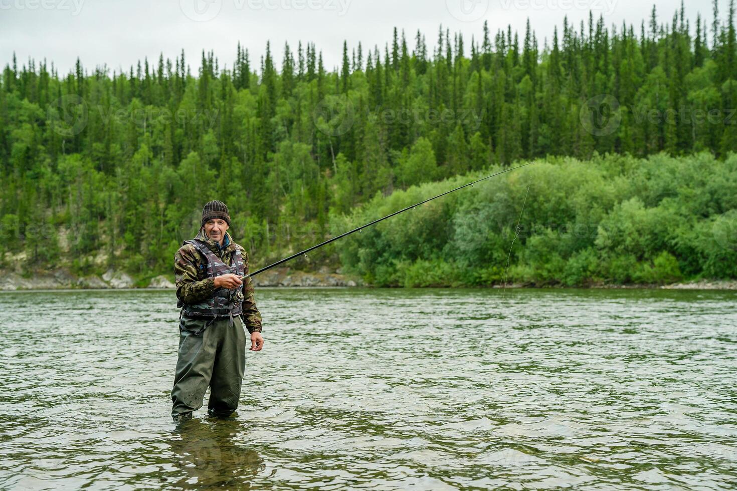 A fisherman stands in a river near a forest with a fishing rod photo