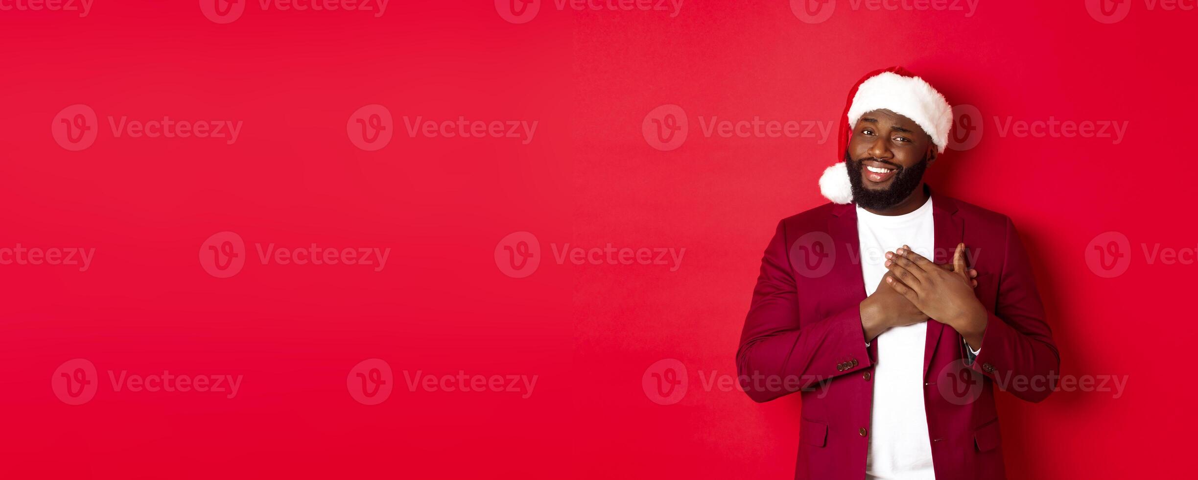 Christmas, party and holidays concept. Grateful african american man in santa hat saying thank you, holding hands on heart and smiling, feeling touched, standing against red background photo