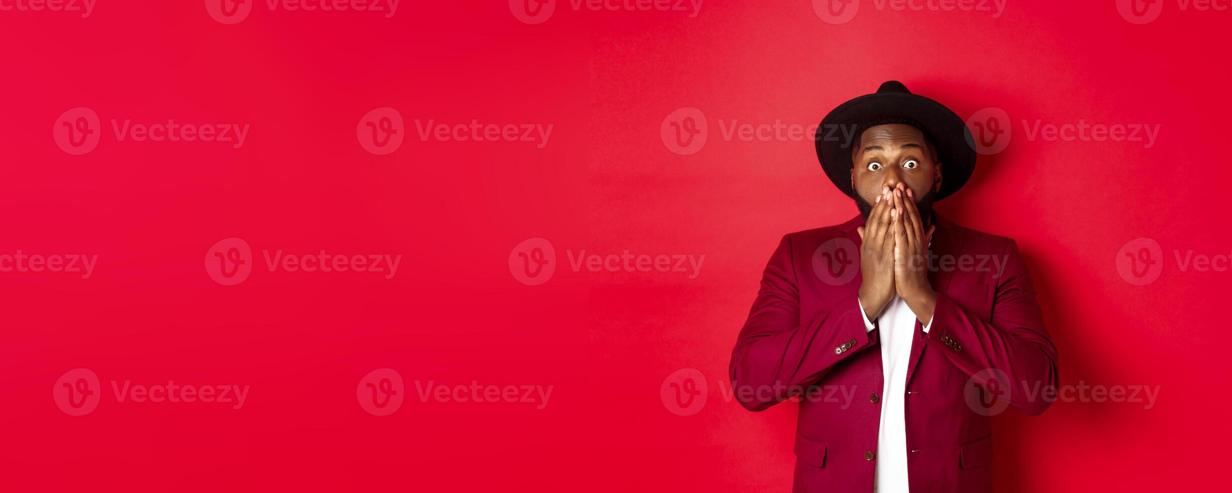 Fashion and party concept. Shocked Black man react on christmas advertisement, gasping and staring at camera with disbelief, standing over red background photo