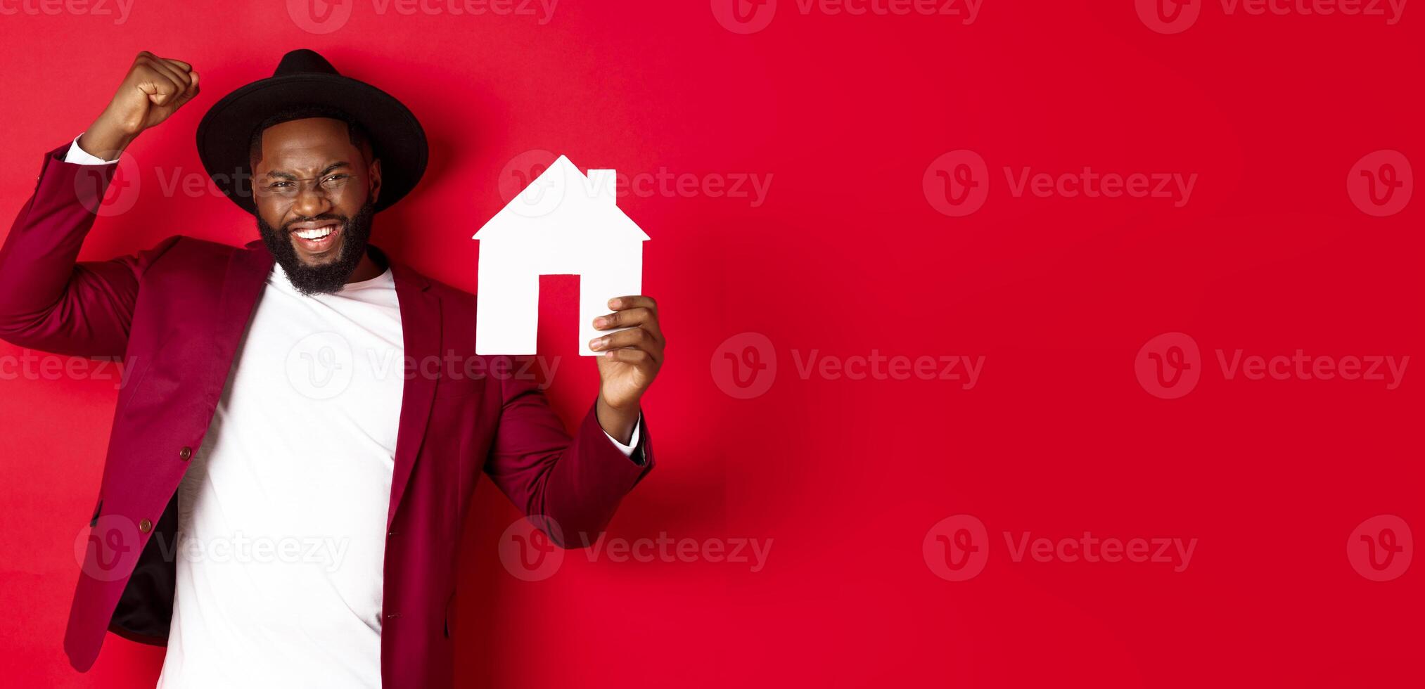 Real estate. Happy Black man celebrating, buying new house, showing paper home model and raising fist up in triumph, standing over red background photo