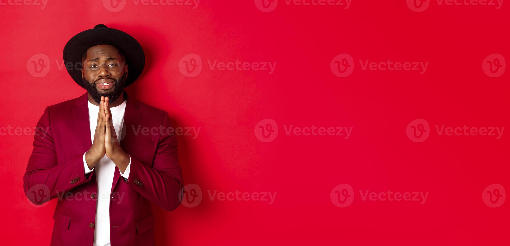Miserable Black man asking for help, begging for favour and looking sad, standing against red background photo