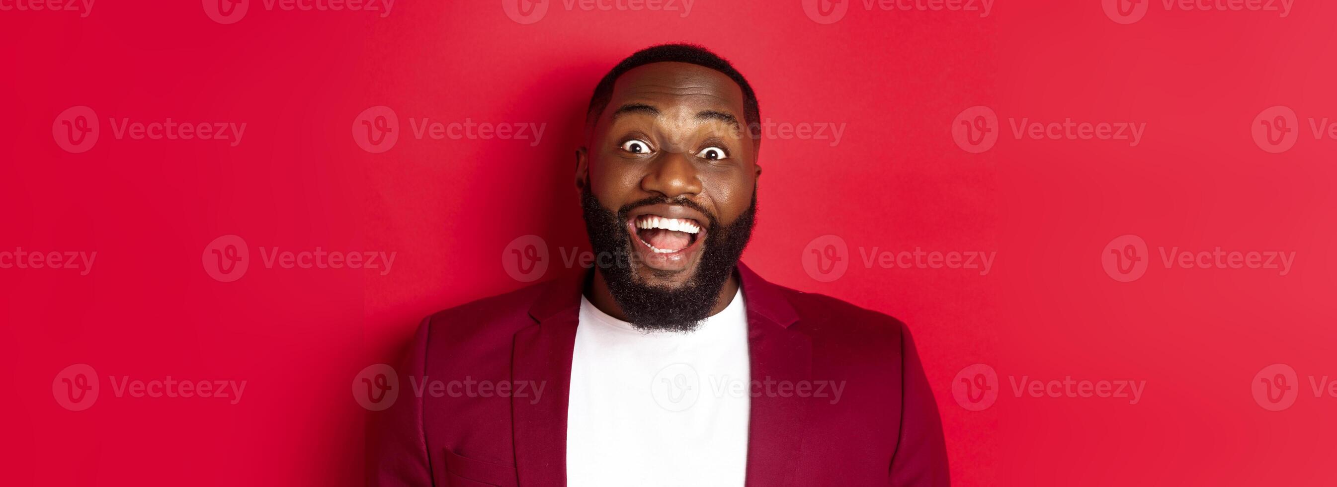 Close-up of african american man with beard staring amazed at camera, smiling and looking excited, standing in blazer against red background photo