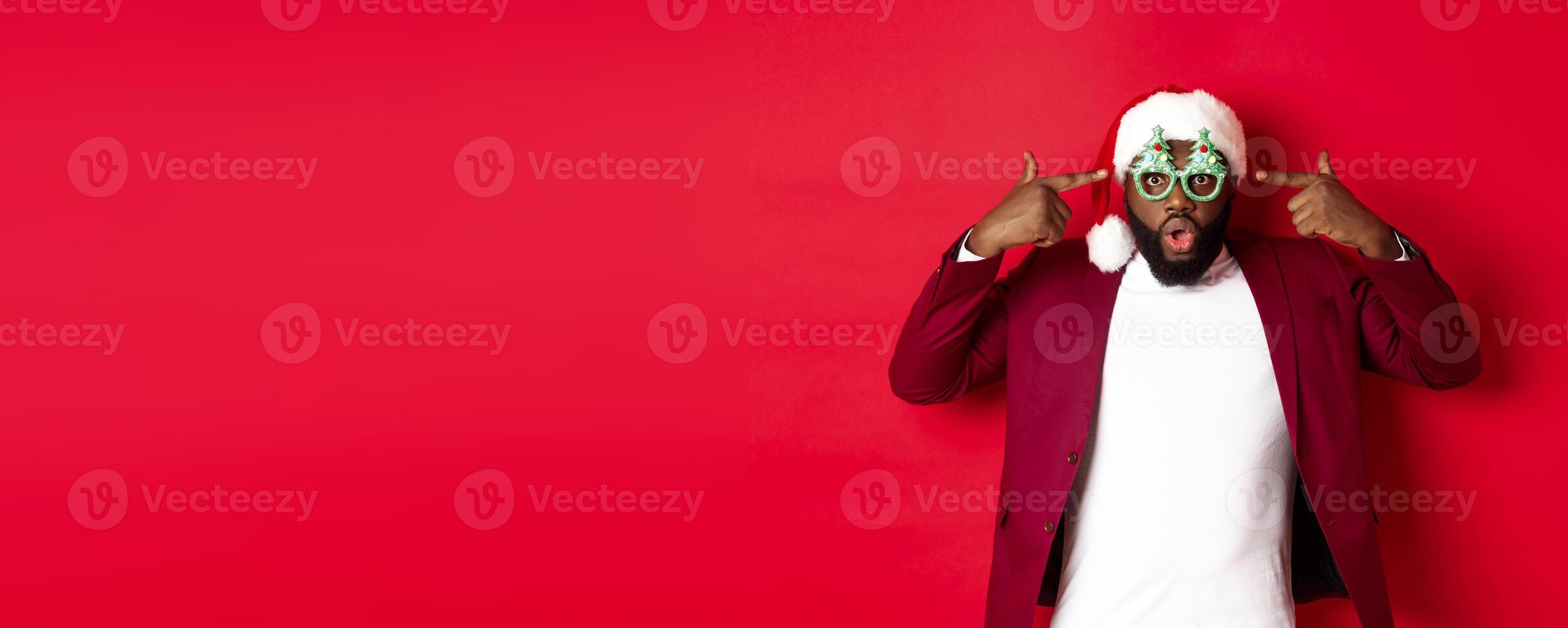 Merry Christmas. Cheerful Black man wearing funny party glasses and santa hat, smiling joyful, celebrating winter holidays, standing over red background photo