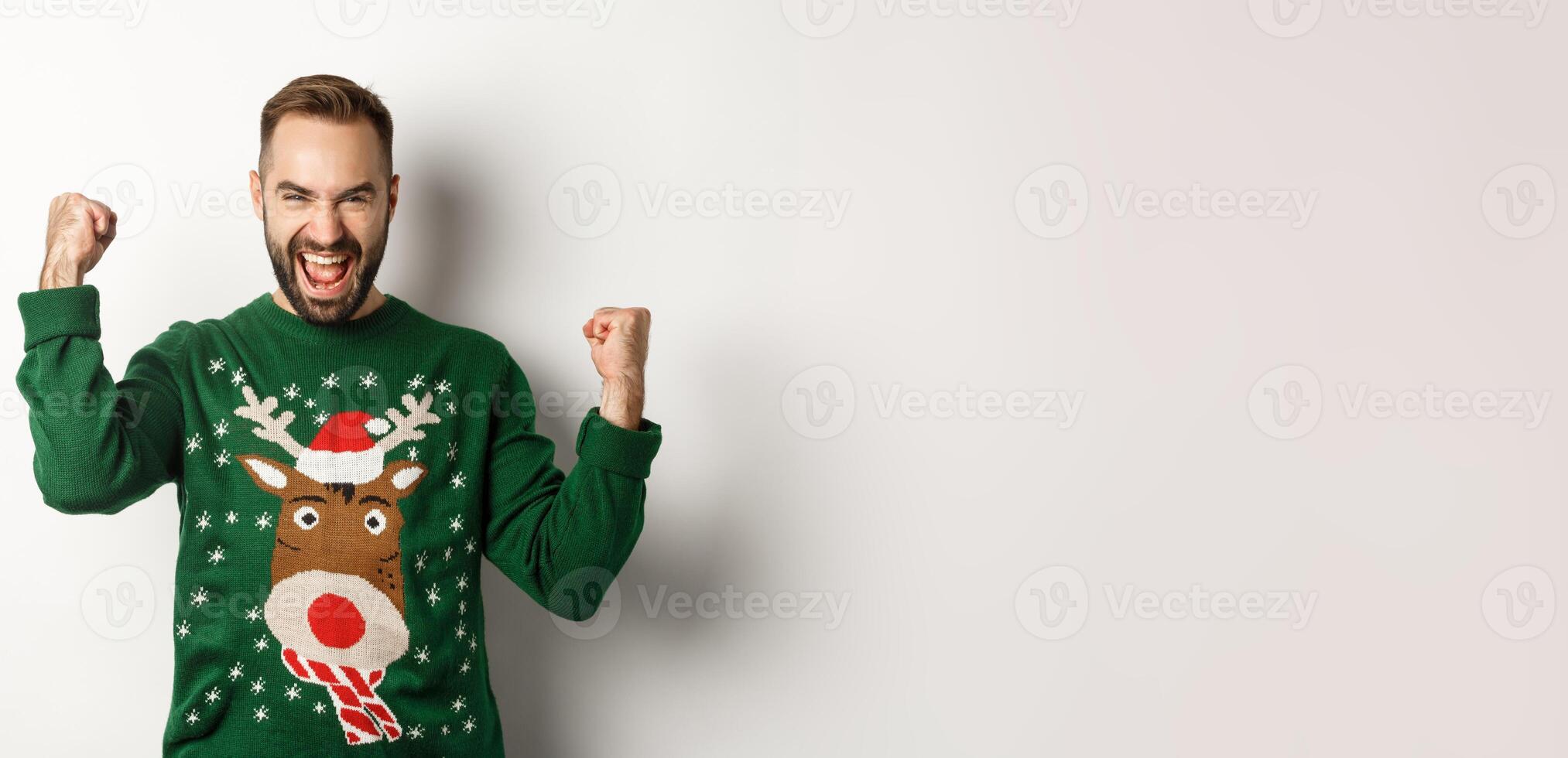 New Year, holidays and celebration. Excited bearded man in Christmas sweater, making fist pumps and shouting for joy, rejoicing and triumphing, white background photo