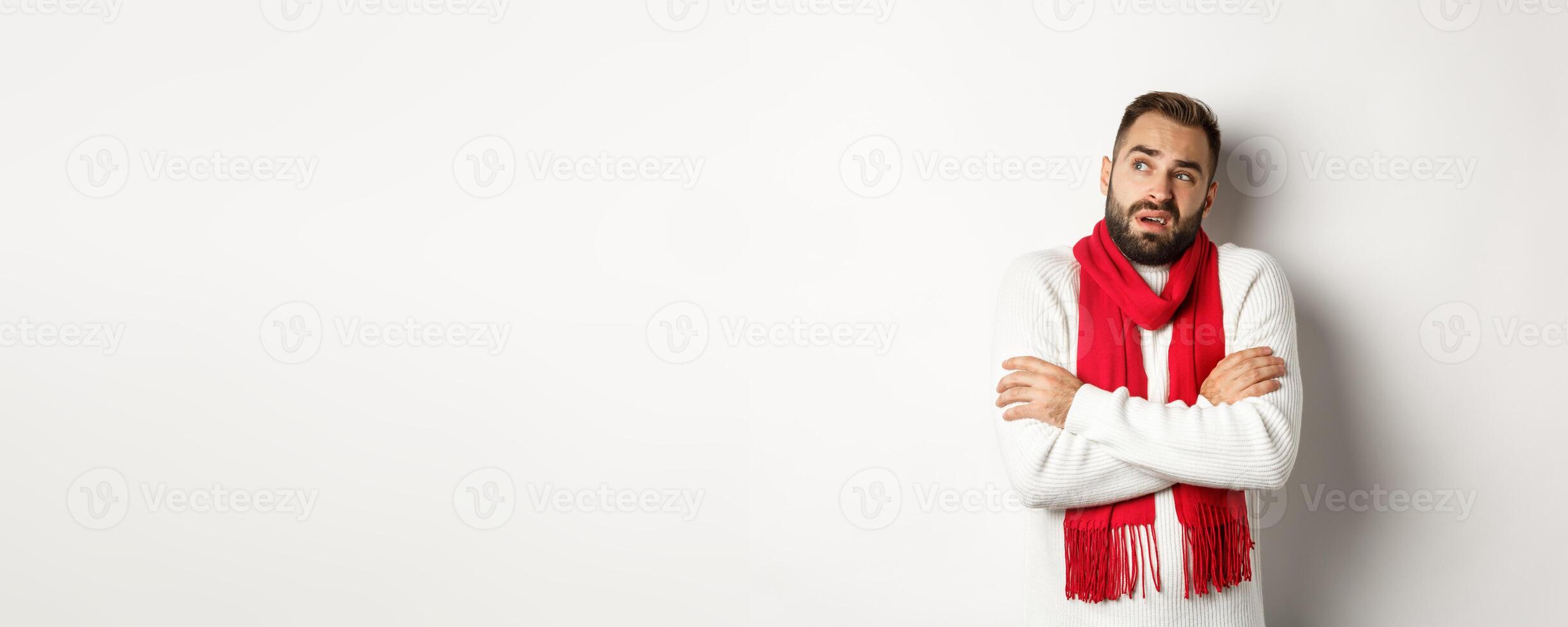 Confused man looking doubtful, standing with hands crossed and feeling cold on winter, wearing christmas sweater with red scarf, standing over white background photo