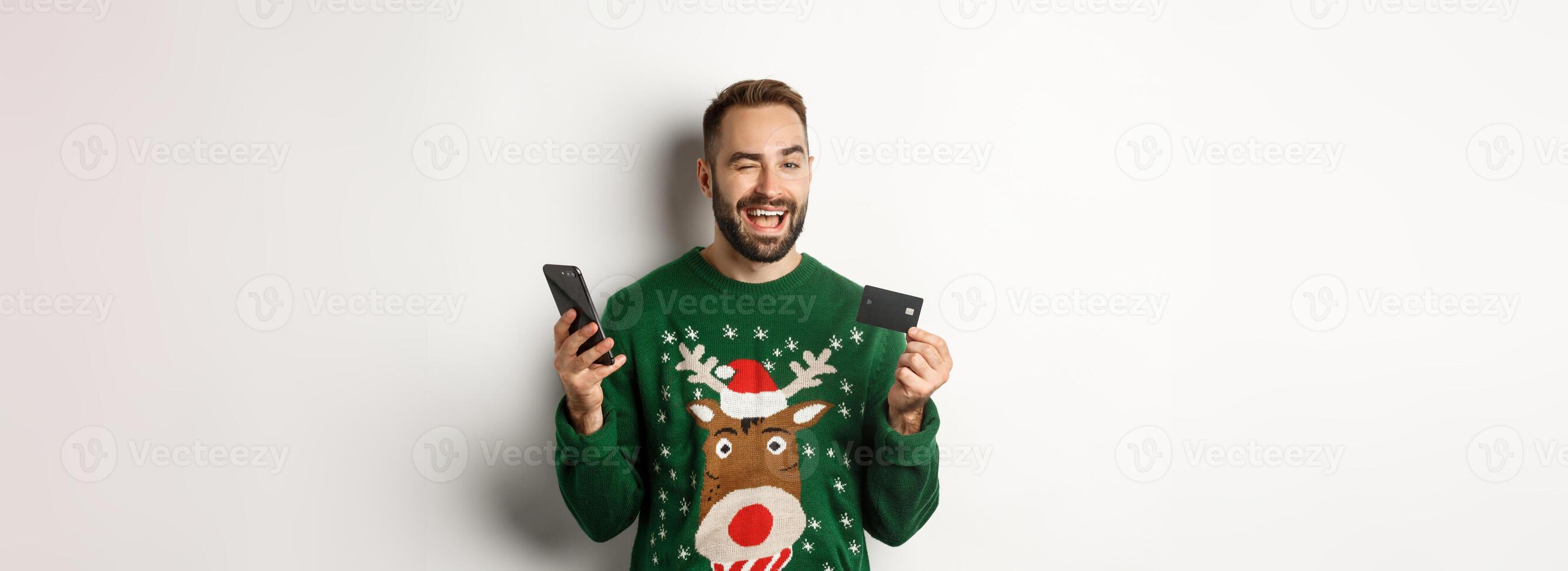 New Year, online shopping and christmas concept. Happy bearded man using mobile phone and credit card, winking at camera, order something in internet, white background photo