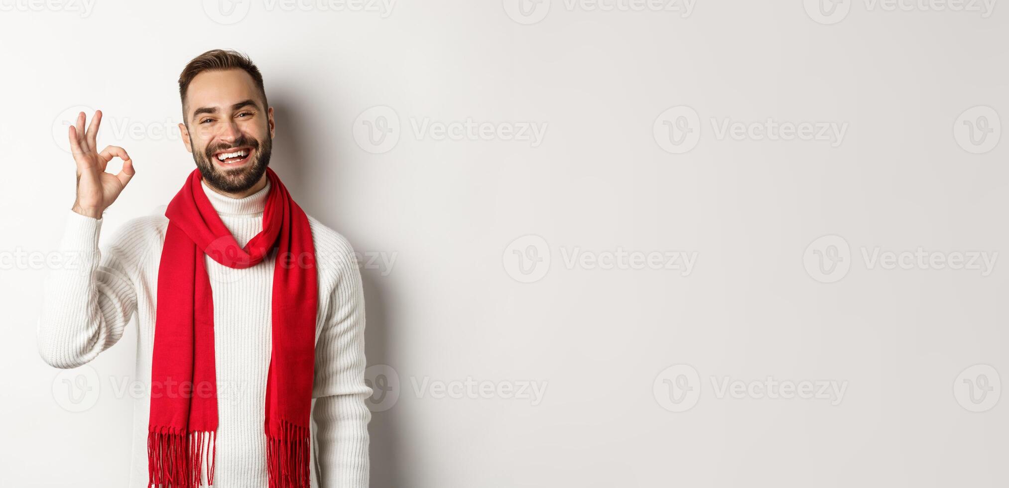 Winter holidays and shopping concept. Satisfied bearded man laughing and showing okay sign, approve and like product, standing in christmas scarf and sweater, white background photo