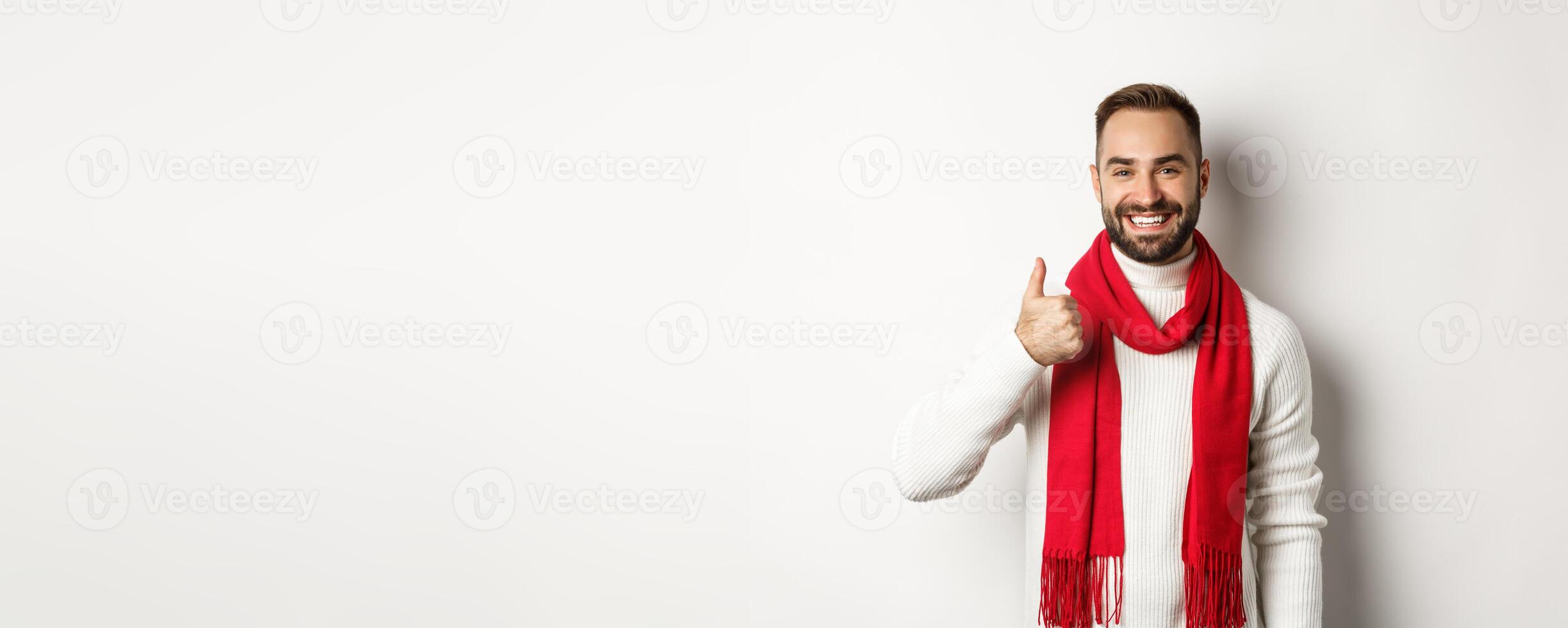 Winter holidays and shopping concept. Confident handsome man showing thumb up, standing in christmas sweater and red scarf, white background photo