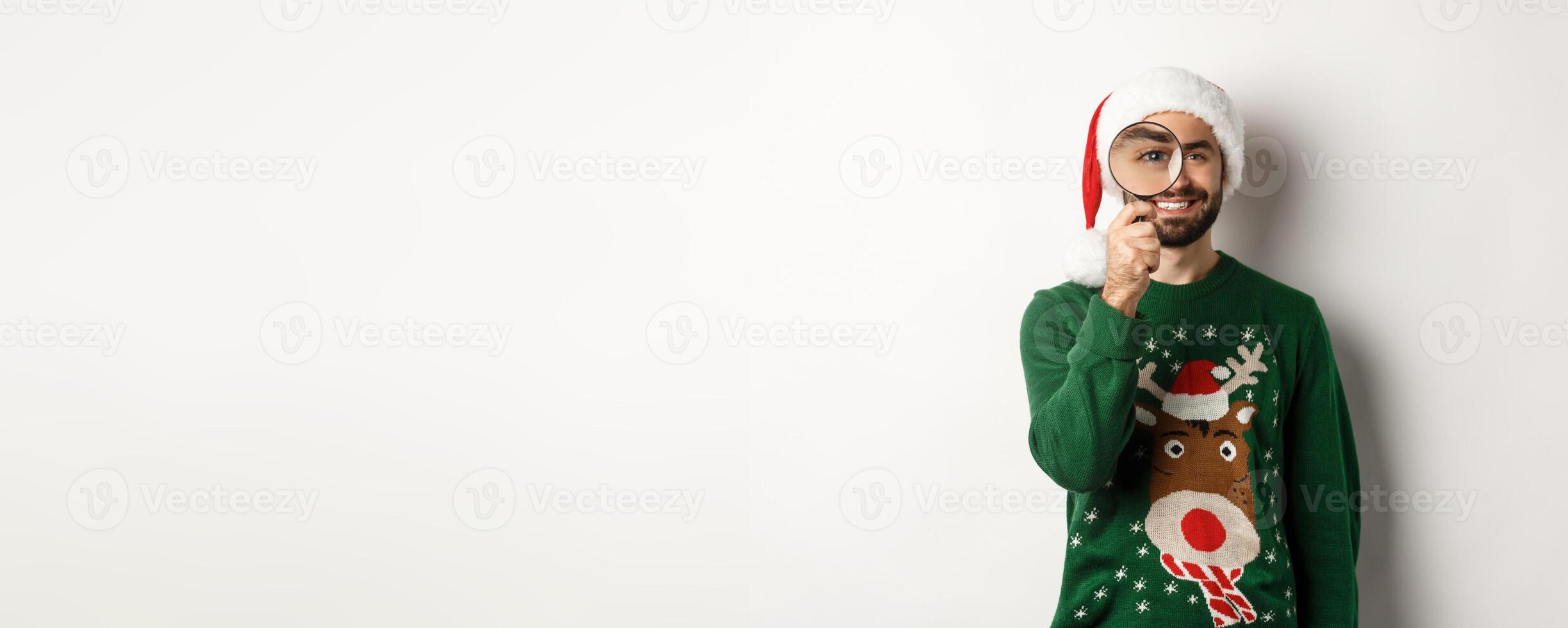 Christmas and holidays concept. Happy bearded man in Santa hat and xmas sweater, looking through magnifying glass and smiling, standing over white background photo