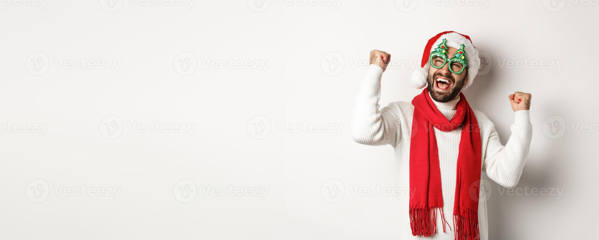 Christmas, New Year and celebration concept. Happy male model in party glasses and santa hat rejoicing, making fist pump and triumphing, standing over white background photo