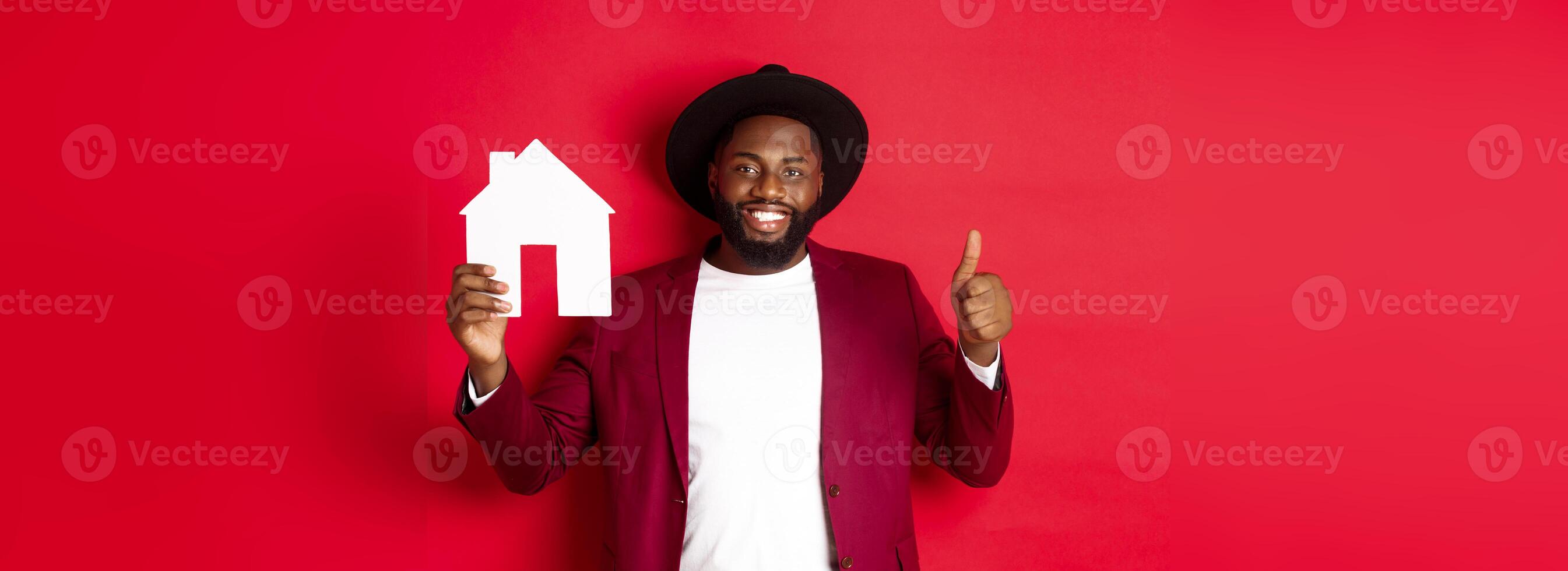 Real estate. Stylish african american bearded man showing home maket, thumb up, recommending agency or apartment complex, standing over red background photo