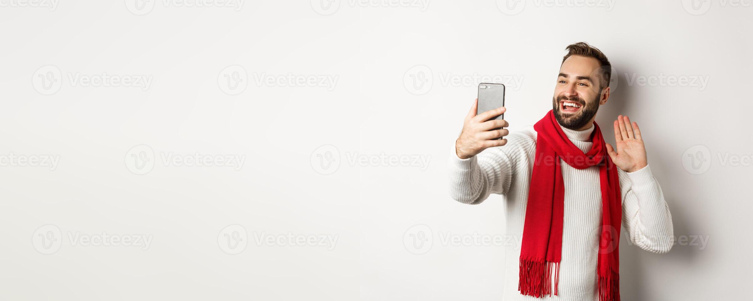 Handsome bearded man calling friends, saying hello and waving hand at mobile phone, standing in sweater and red scarf against white background photo