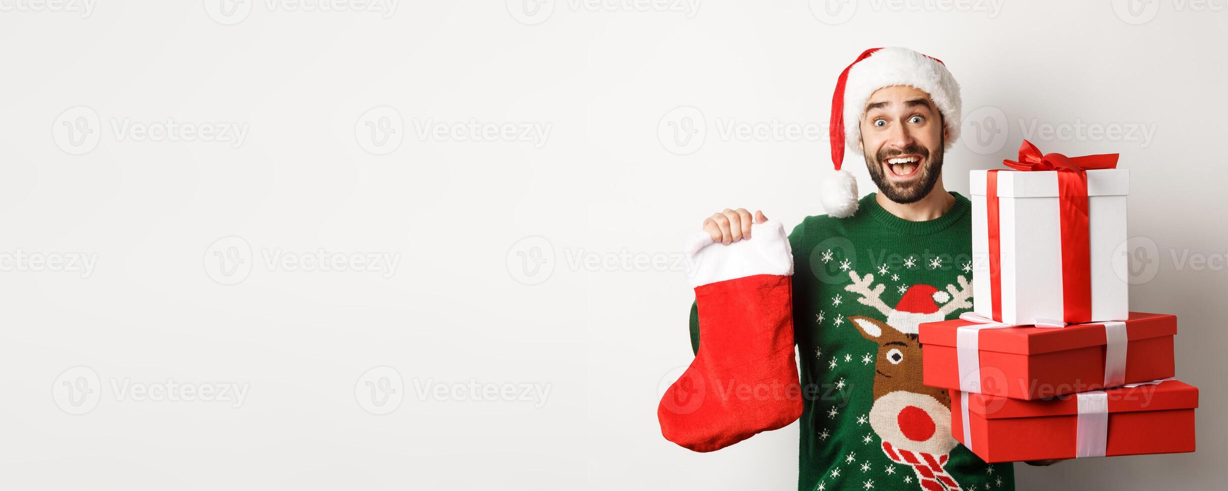 Xmas and winter holidays concept. Excited man holding christmas sock and gift boxes, celebrating New Year, standing over white background photo