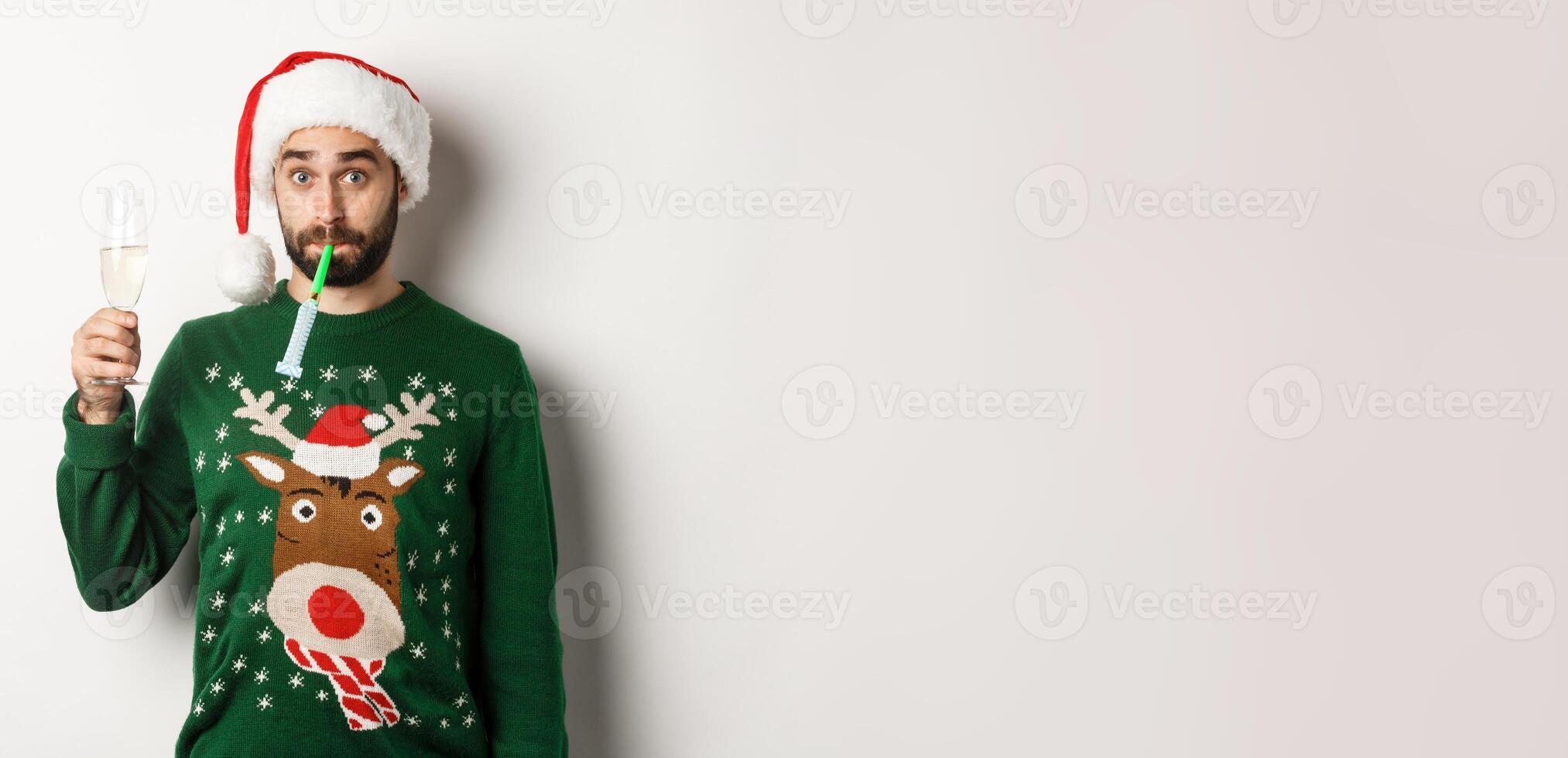 Christmas and holidays concept. Funny guy in Santa hat blowing a party whistle, drinking champagne, standing over white background photo