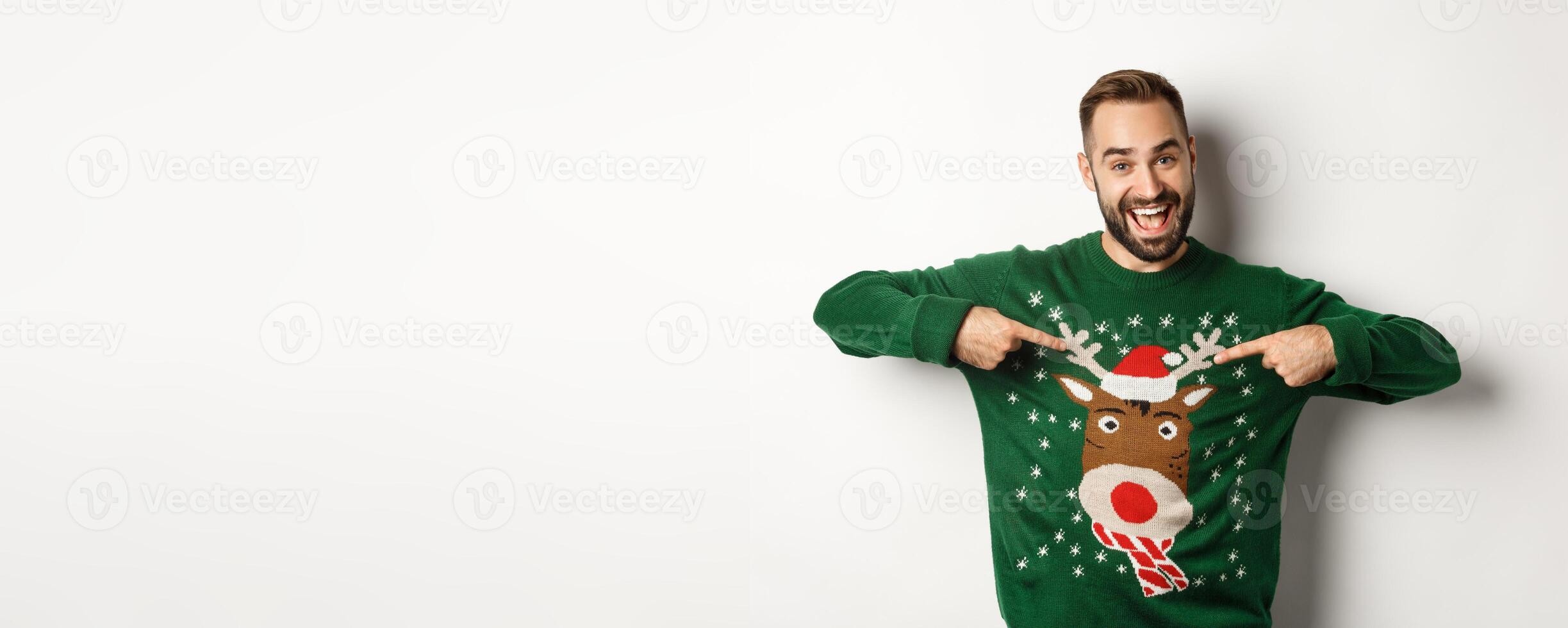 New year celebration and winter holidays concept. Cheerful man pointing at his funny Christmas sweater and smiling, white background photo