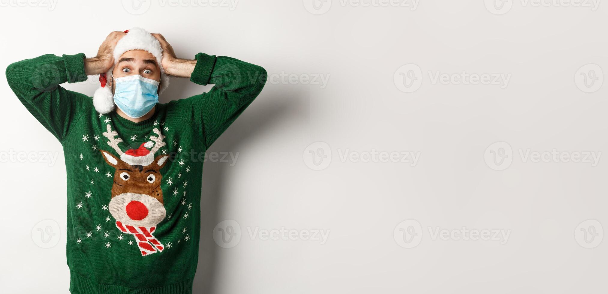 Concept of covid-19 and Christmas holidays. Excited and super happy man in medical mask and Santa hat looking amazed, white background photo
