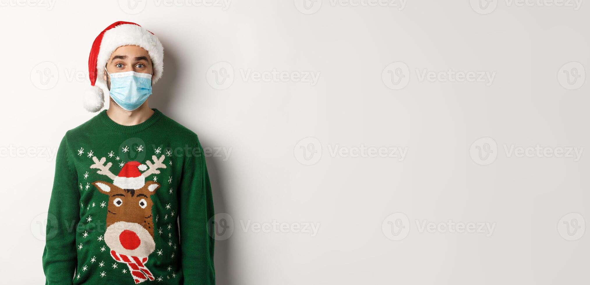 Christmas during pandemic, covid-19 concept. Young man in Santa hat and face mask celebrating New Year party with preventive measures, white background photo