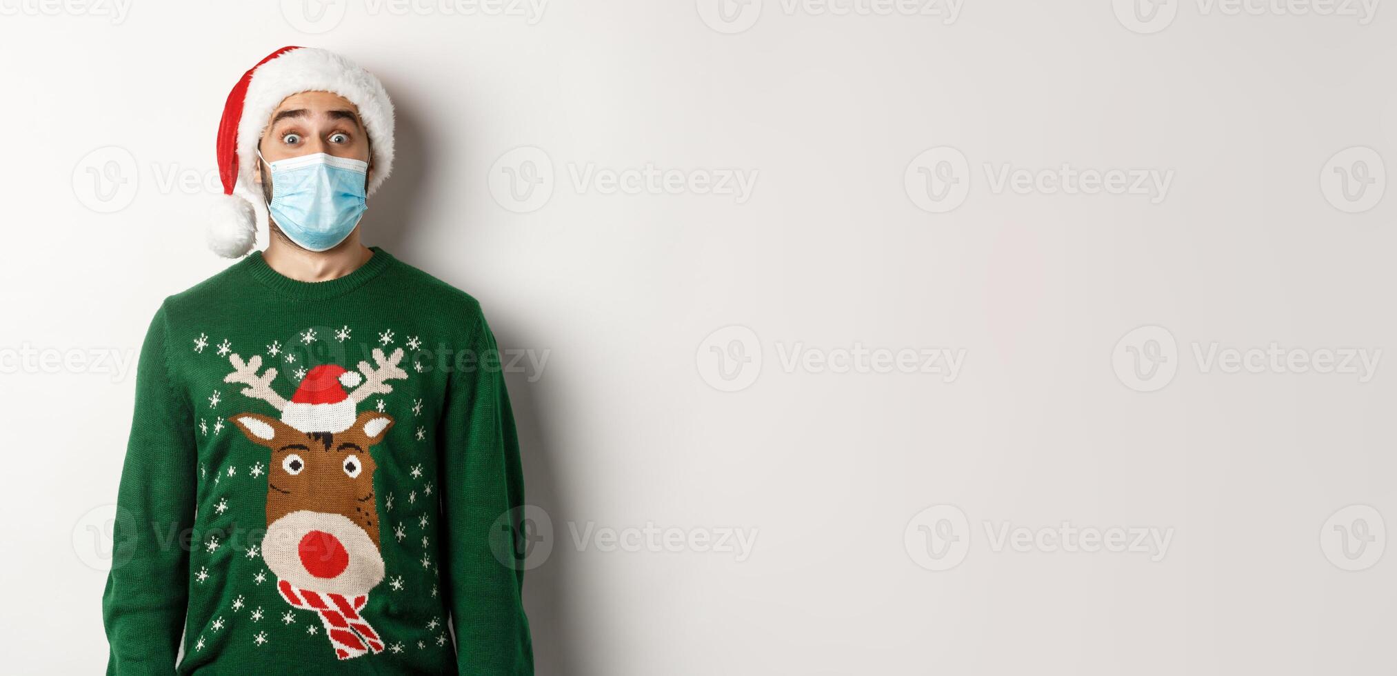 Christmas during pandemic, covid-19 concept. Surprised guy in medical mask, Santa hat and sweater celebrating New Year party, standing over white background photo